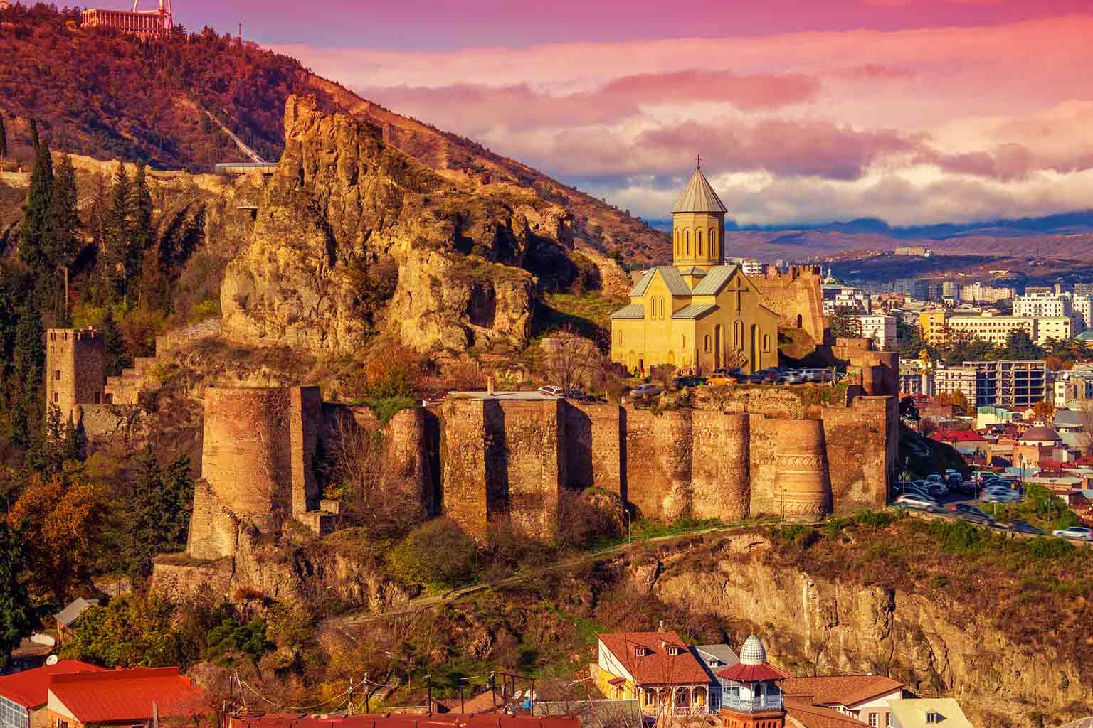 Tbilisi cityscape at sunset featuring ancient stone fortress ruins, yellow Armenian church with cross dome, and modern city b